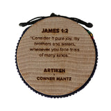 A round wooden prayer wheel with a black cord, featuring engraved text including 'JAMES 1:2' and 'CONNER MANTZ ARTIKEN'.
